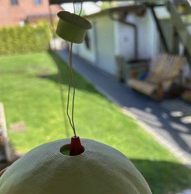 Eine grüne Kugel mit einer roten und einer grünen Kappe, im Vordergrund hängen. Im Hintergrund eine Gartenlandschaft.