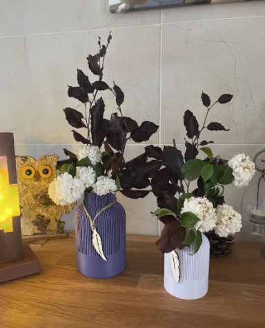 Zwei Vasen mit weißen Blumen und dunkelgrünem Blattwerk auf einem Tisch.