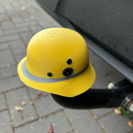 Gelber Sicherheitshelm mit gräulicher Band und Augen, auf einem Autoanhänger montiert.