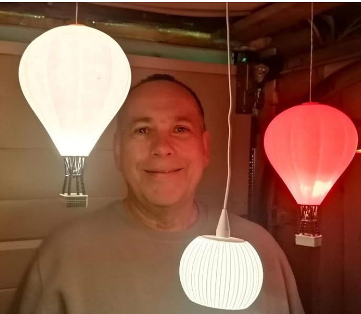 Mann lächelt zwischen drei hängenden Lampen in Form von Heißluftballons.
