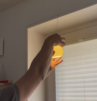 Eine Hand hebt eine Lampe, die an einem Fenster mit Jalousien hängt.